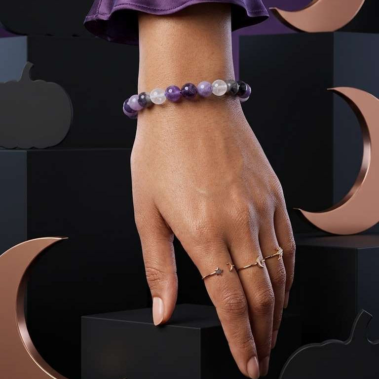 A person's hand wearing a beaded bracelet with purple, white, and gray beads, styled with slim gold rings, set against a dark background with decorative gold crescent moon and pumpkin shapes.